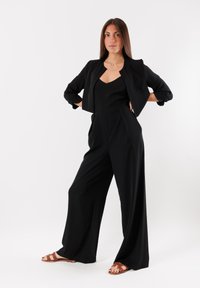 Tuta nera con gamba larga e scollatura a V, abbinata a un blazer nero corto. La modella indossa sandali marroni con cinturini e si trova su uno sfondo neutro.