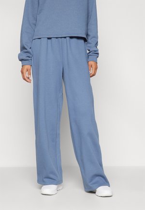 Person trägt lockere blaue Jogginghose und passendes Sweatshirt mit weißen Turnschuhen, steht vor einem schlichten weißen Hintergrund.