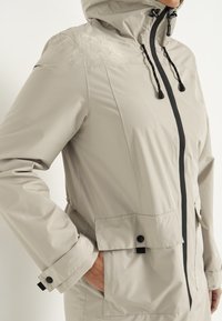 Leichte beige Regenjacke mit durchgehendem Reißverschluss, verstellbare Kapuze, Seitentaschen und schwarzen Akzenten an Reißverschluss und Ärmeln. Wasserdichtes Material.