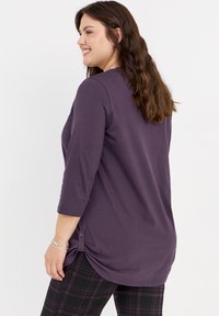Lila Bluse mit drei Viertelärmeln und abgerundetem Saum, mit Knopfdetail an der Seite, kombiniert mit karierter Hose. Weicher Stoff.