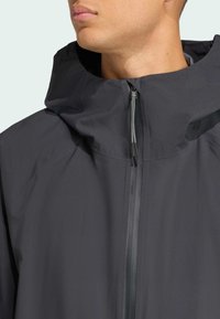 Mørkegrå vandtæt jakke med høj krave, justerbar hætte og fuld længde frontlynlås. Har et slankt, minimalistisk design.