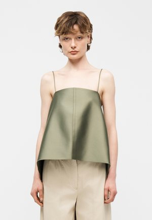 Ung person med kort, bølget hår iført en olivengrøn ærmeløs top med tynde stropper og beige bukser med vide ben, stående mod en hvid baggrund.