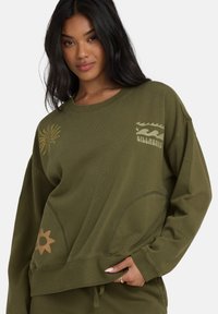 Felpa verde oliva con scollatura rotonda, vestibilità ampia e maniche lunghe. Presenta motivi stampati di sole e onde in toni beige a contrasto.