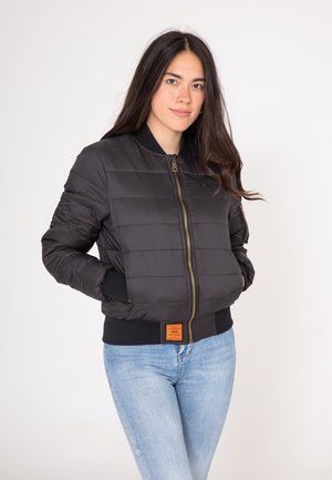 Giubbotto invernale nero con texture a cerniera orizzontale, zip dorata, polsini a costine e un'etichetta arancione sull'orlo. Indossato con jeans blu.