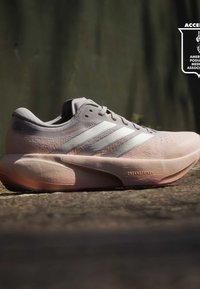 Ανοιχτό ροζ παπούτσι για τρέξιμο Adidas με λευκές ρίγες στο πεζοδρόμιο, πλευρική όψη που δείχνει τις λεπτομέρειες κειμένου "Supernova" και "Dreamstrike".