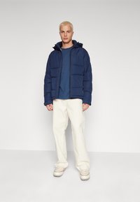 Giubbotto imbottito blu navy con cappuccio, caratterizzato da quadrati trapuntati e chiusura con zip, indossato sopra una maglia blu. Abbinato a pantaloni crema e sneaker bianche.