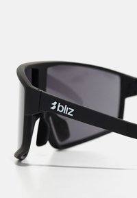Czarne sportowe okulary przeciwsłoneczne o matowym wykończeniu, zakrzywionym kształcie i szarych szkiełkach. Zawierają białe logo marki na oprawce.
