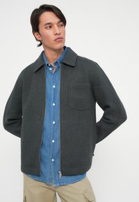 Mørkegrå zip-jakke med flat krage og brystlomme, lagd over en lys blå skjorte med knapper. Glatt ullstruktur.