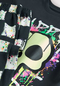 T-shirt di cotone nero con una grande grafica di teschio verde e un design colorato a spruzzi. Presenta un tessuto coordinato a motivo alla cintura.