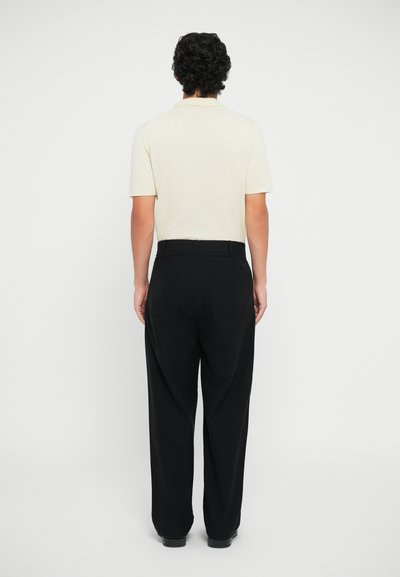 Polo de manga corta crema, pantalones negros, vista trasera. Los pantalones tienen un corte relajado y bolsillos. Textura suave con un detalle mínimo.