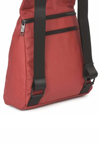 Mochila roja de nailon con textura acolchada, dos bolsillos con cremallera y correas negras ajustables. Presenta una forma rectangular y detalles de herrajes resistentes.