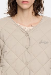 Quilted beige jacket with a rounded neckline, featuring two snap buttons and embroidered logo on the front. Smooth, soft fabric texture.