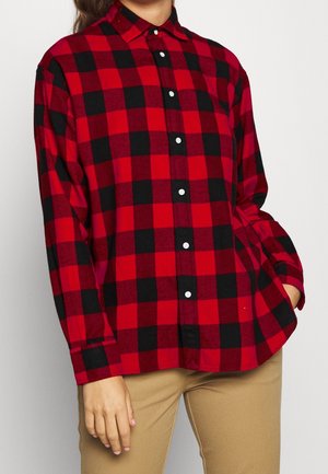 Person wearing a red and black checkered button-up shirt with long sleeves paired with light brown pants, hand resting in pocket.