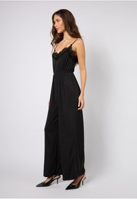 Svart jumpsuit laget av mykt stoff, med blonderkantet utringning, vide ben og tynne stropper. Kombinert med stilige svarte hæler.