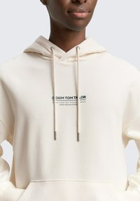 Hoodie crème en tissu doux, avec une poche avant, un col à cordons et une marque imprimée en bleu foncé.