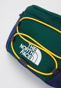 Zielono-granatowa torba The North Face z żółtymi elastycznymi sznurkami z przodu i zamkiem błyskawicznym na górze.