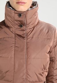 Chaqueta marrón acolchada con cuello alto, que cuenta con dos botones y un forro suave gris. Fabricada con un tejido brillante y liso.