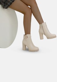 Botas de tobillo beige con un tacón texturizado, detalle de hebilla y cremallera lateral. Presentan una suela plataforma gruesa con un diseño estriado.