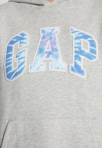 Sudadera con capucha gris con grandes letras "GAP" en un patrón de teñido anudado azul con costuras blancas. Incluye un bolsillo frontal y tela suave y texturizada.