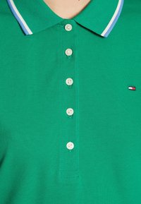 Zielona koszulka polo z prążkowanym kołnierzykiem w niebieskie i białe paski. Posiada pięć guzików oraz małe logo w prawym dolnym rogu.