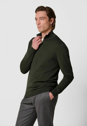 Uomo dalla pelle chiara e capelli castani che indossa un maglione verde scuro con cerniera e pantaloni grigi, posando con una mano vicino al colletto e l'altra in tasca.