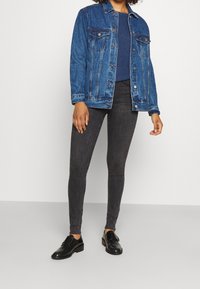 Veste en jean bleu moyen, dotée de poches poitrine et de fermetures à boutons, associée à un jean noir slim et des chaussures noires.