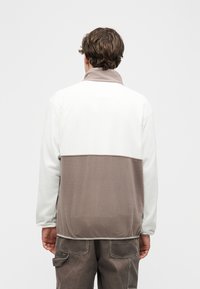 Άνδρας που φορά λευκό και γκρι μπουφάν φλις και σκούρο γκρι παντελόνι εργασίας, στέκεται με την πλάτη προς τα εμπρός σε απλό ανοιχτό φόντο.