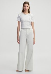 T-shirt blanc à manches courtes avec un motif subtil et un détail en chaîne argentée, associé à un pantalon large taille haute gris clair.