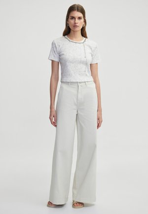T-shirt blanc à manches courtes avec un motif subtil et un détail en chaîne argentée, associé à un pantalon large taille haute gris clair.