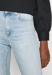 Jeans skinny de mezclilla azul claro con cintura media, que cuentan con bolsillos frontales y un leve desgastado, combinados con una camiseta negra de manga larga.