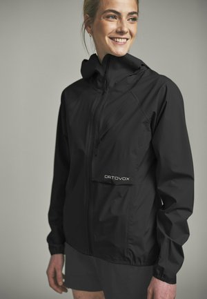 Junge Frau lächelnd, trägt eine schwarze Ortovox Outdoor-Jacke mit einer Reißverschlusstasche auf der Brust, steht vor neutralem Hintergrund.