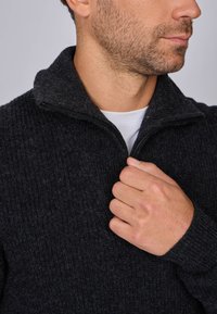 Gilet zippé gris foncé avec un col haut, motif en maille texturée et poignets côtelés, porté partiellement zippé au cou.