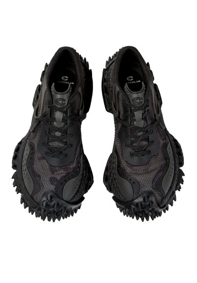 Sorte high-tech sneakers med teksturerede, piggede såler og mesh-detaljer, set ovenfra med synlige snørebånd og brandlogo indeni.