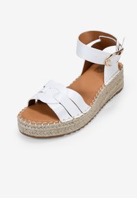 Sandalia cuña de esparto blanca de cuero con diseño de correas cruzadas, correa para el tobillo y hebilla dorada. La plataforma envuelta en yute añade textura.
