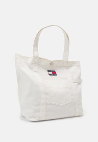 Vit tygväska med beige sömmar, med en framficka och en logopatch. Två robusta handtag för bärande.