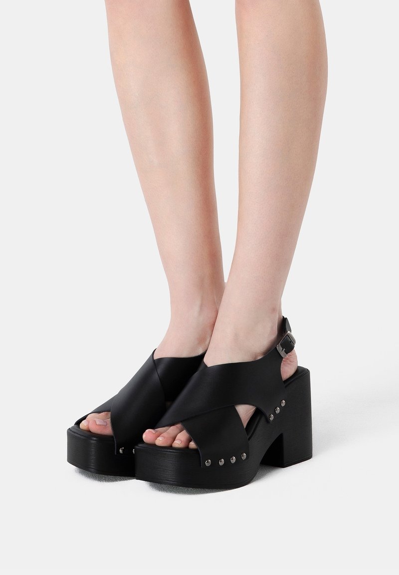 Personne portant des sandales plateformes noires avec des talons épais et des brides croisées maintenues par une boucle sur fond blanc.