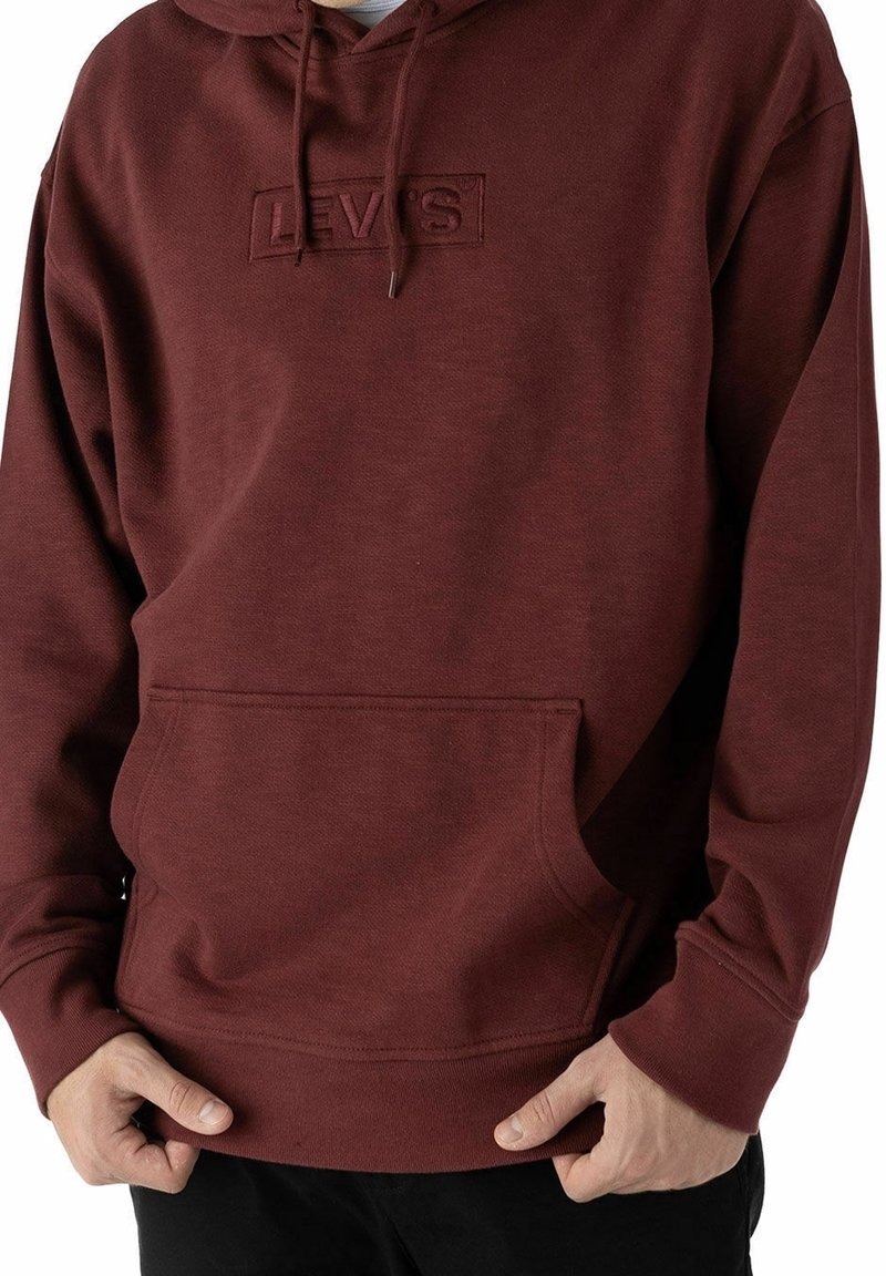 Homme portant un sweat à capuche Levi's bordeaux avec poche avant et cordon de serrage à la capuche, les mains tenant l'ourlet inférieur.