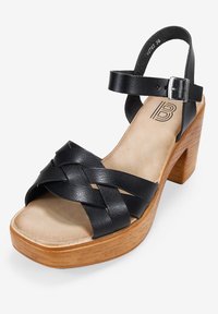 Sandali con tacco in pelle nera, con cinturini intrecciati, punta aperta e cinturino alla caviglia. Tacco a blocco in legno e soletta morbida color beige.