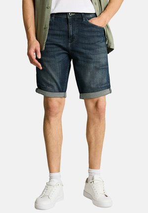 Man wearing rolled dark denim shorts, white socks, white sneakers, white t-shirt, and an open green shirt.