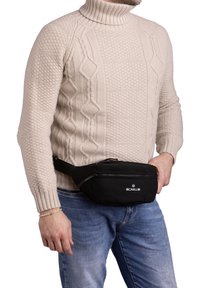 Marsupio nero in tessuto resistente, con tasca con zip e tracolla regolabile, indossato sopra un maglione beige a trecce.