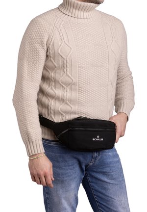 Marsupio nero in tessuto resistente, con tasca con zip e tracolla regolabile, indossato sopra un maglione beige a trecce.