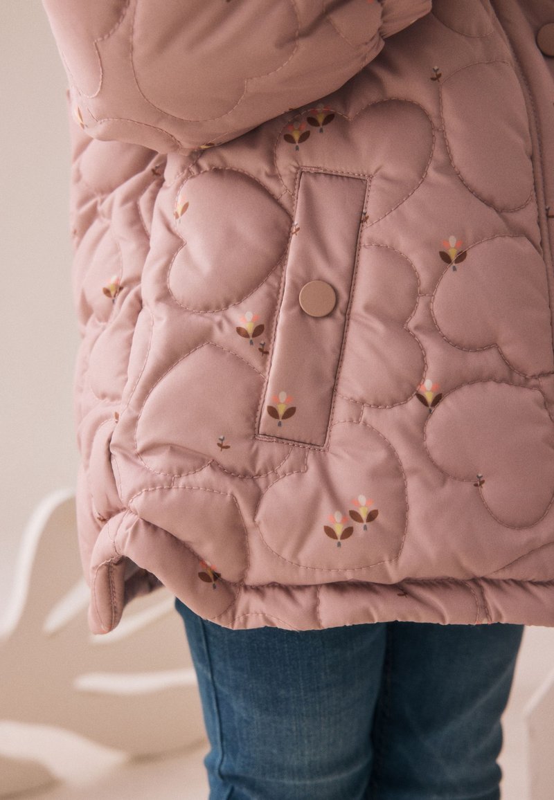 Veste matelassée rose avec motifs en forme de cœur et petits imprimés floraux, avec une poche à pression, portée sur un jean bleu.