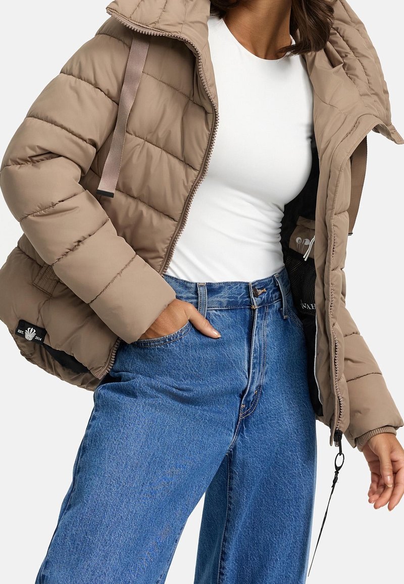 Beige gewatteerde jas met een doorgestikt design, voorzien van een ritssluiting, grijze accentbanden en een zak met een mesh voering. Gecombineerd met een blauwe spijkerbroek.