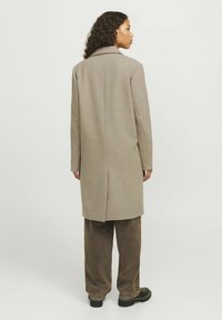 Abrigo largo gris hecho de una mezcla de lana, con solapa en pico, corte recto y una costura central en la parte posterior, combinado con pantalones anchos beiges y zapatos negros.