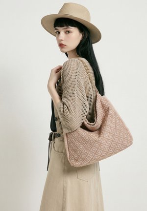 Vrouw met lang zwart haar die een beige hoed draagt, een opengewerkte cardigan en een grote, gestructureerde roze schoudertas met een diamantpatroon.