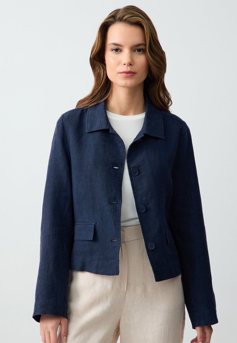 Marineblauwe linnen blazer met een kraaghalslijn, voorknopsluiting en twee klepzakken, gecombineerd met een witte top en lichtbeige broek.