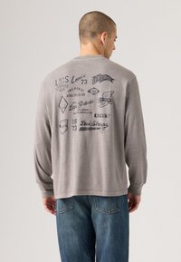Grå langermet skjorte med trykte svarte grafikker, som har logoer og tekst relatert til Levi's. Enkel rund hals og rett kant.