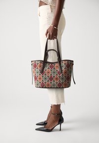 Textile tote bag with a geometric pattern in red, green, and orange, contrasting brown leather handles. Structured shape and zipper detail.