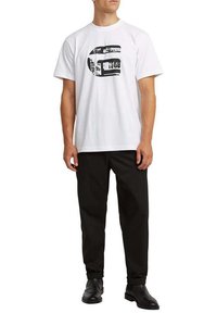 Hombre con una camiseta blanca de manga corta con un logo gráfico negro, pantalones negros y botas negras, de pie frente a un fondo liso.