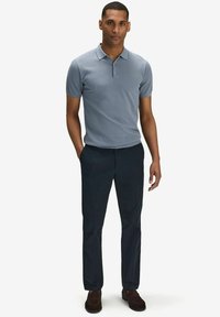 Ljusblå kortärmad poloströja med ett texturerat mönster, i kombination med mörka byxor. Modellen står med händerna i fickorna och har ett neutralt uttryck.